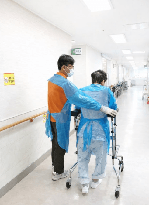 광주 에스웰 요양병원 CRE 격리 환자가 1회용 가운을 입은 이송 선생님과 함께 워커를 이용해 복도 보행 재활을 하는 모습